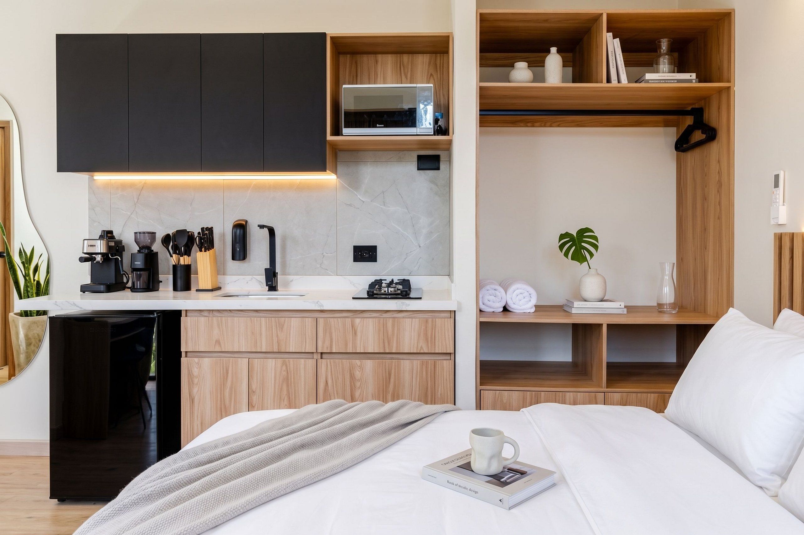 Un dormitorio moderno con cama, una mesita de noche con un libro y una taza, una zona de cocina con cafetera, y un estante de madera con libros y una planta.