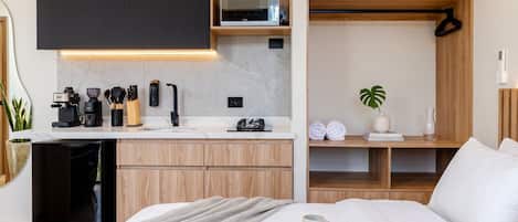 A modern bedroom with a bed, a bedside table with a book and a mug, a kitchen area with a coffee machine, and a wooden shelf with books and a plant.