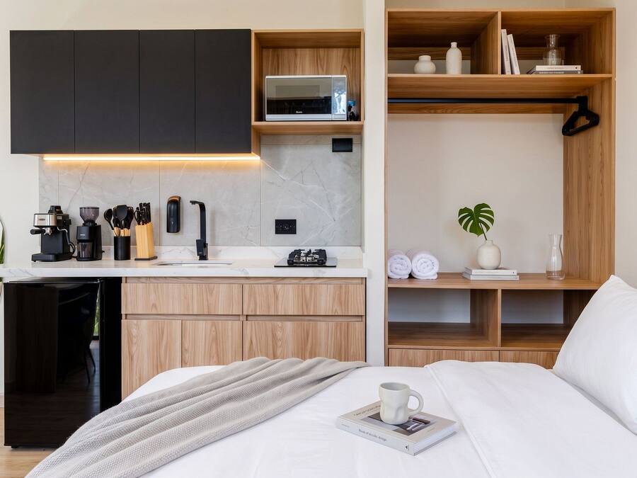 Un dormitorio moderno con cama, una mesita de noche con un libro y una taza, una zona de cocina con cafetera, y un estante de madera con libros y una planta.