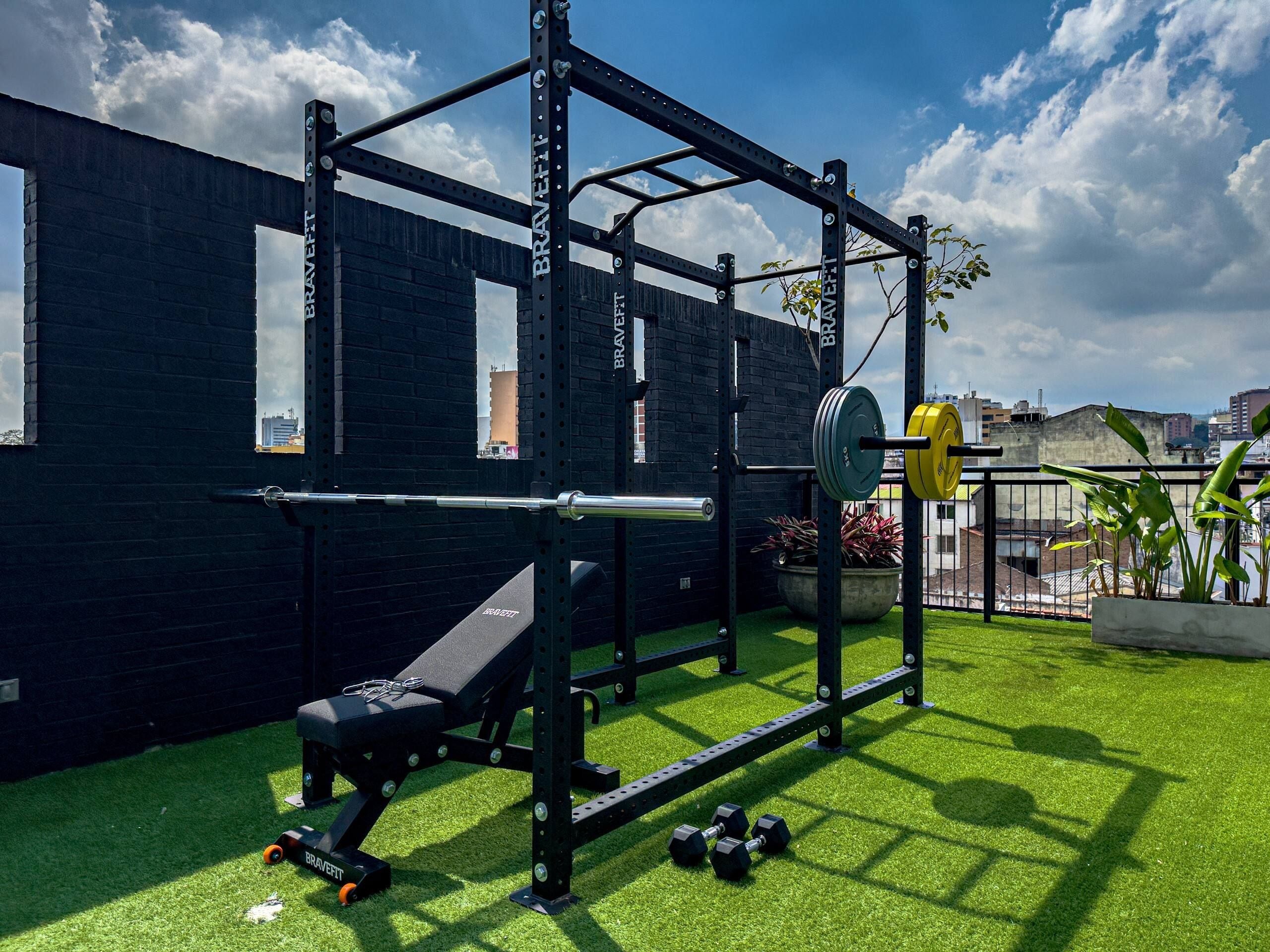 A gym setup with a squat rack, bench, and weights, located outdoors with a brick wall and greenery.
