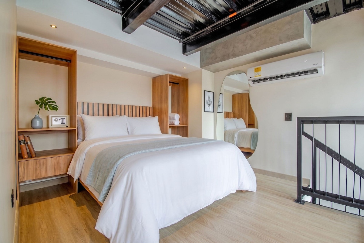 A modern bedroom with a large bed, a wooden headboard, a bedside table with a lamp, a plant, and a framed picture.