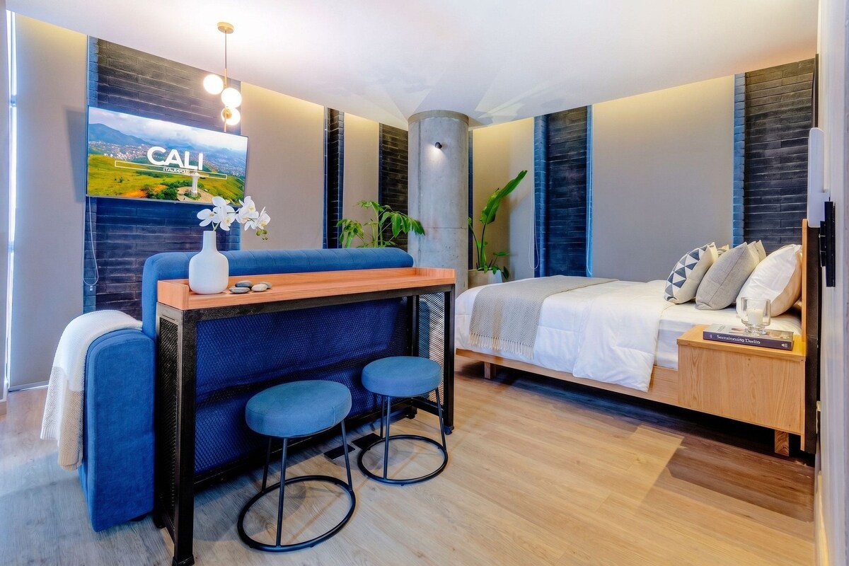 A modern bedroom with a bed, a desk with two chairs, a large window, and a wall-mounted TV.
