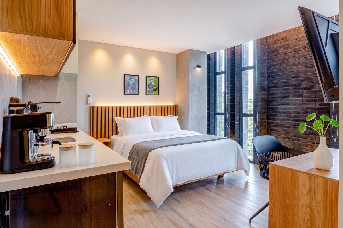 A modern hotel room with a bed, bedside table, coffee maker, and a view of the outdoors.