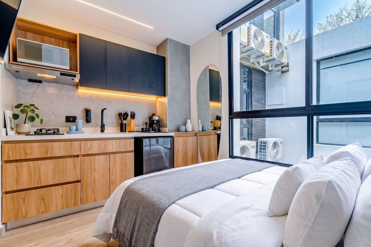 A modern bedroom with a large bed, a kitchen area with wooden cabinets, and a view of the outdoors through large windows.