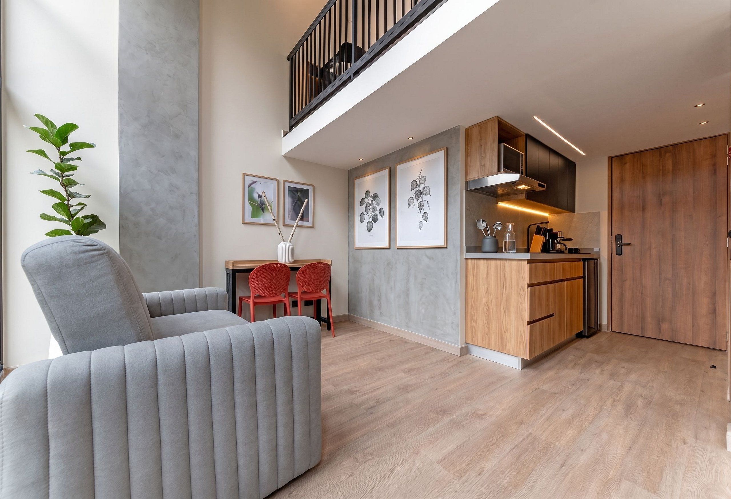 A modern living space with a grey sofa, a dining table with two chairs, a kitchen area with wooden cabinets, and a staircase leading to an upper level.