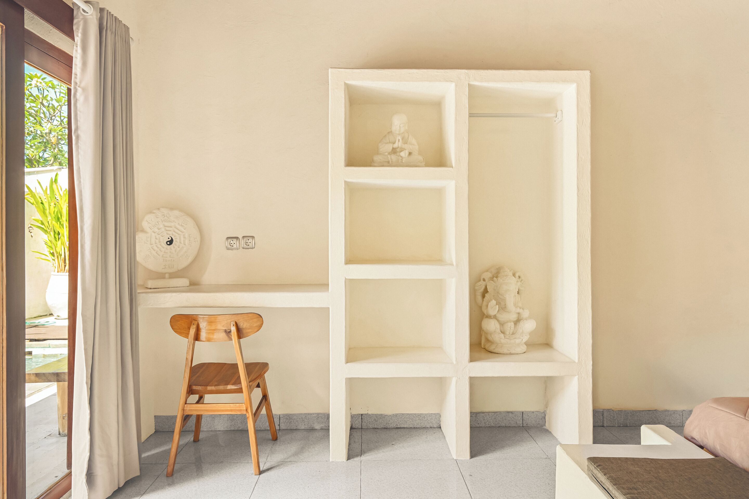 A room with a desk, a chair, a shelf with statues, and a view of greenery outside.