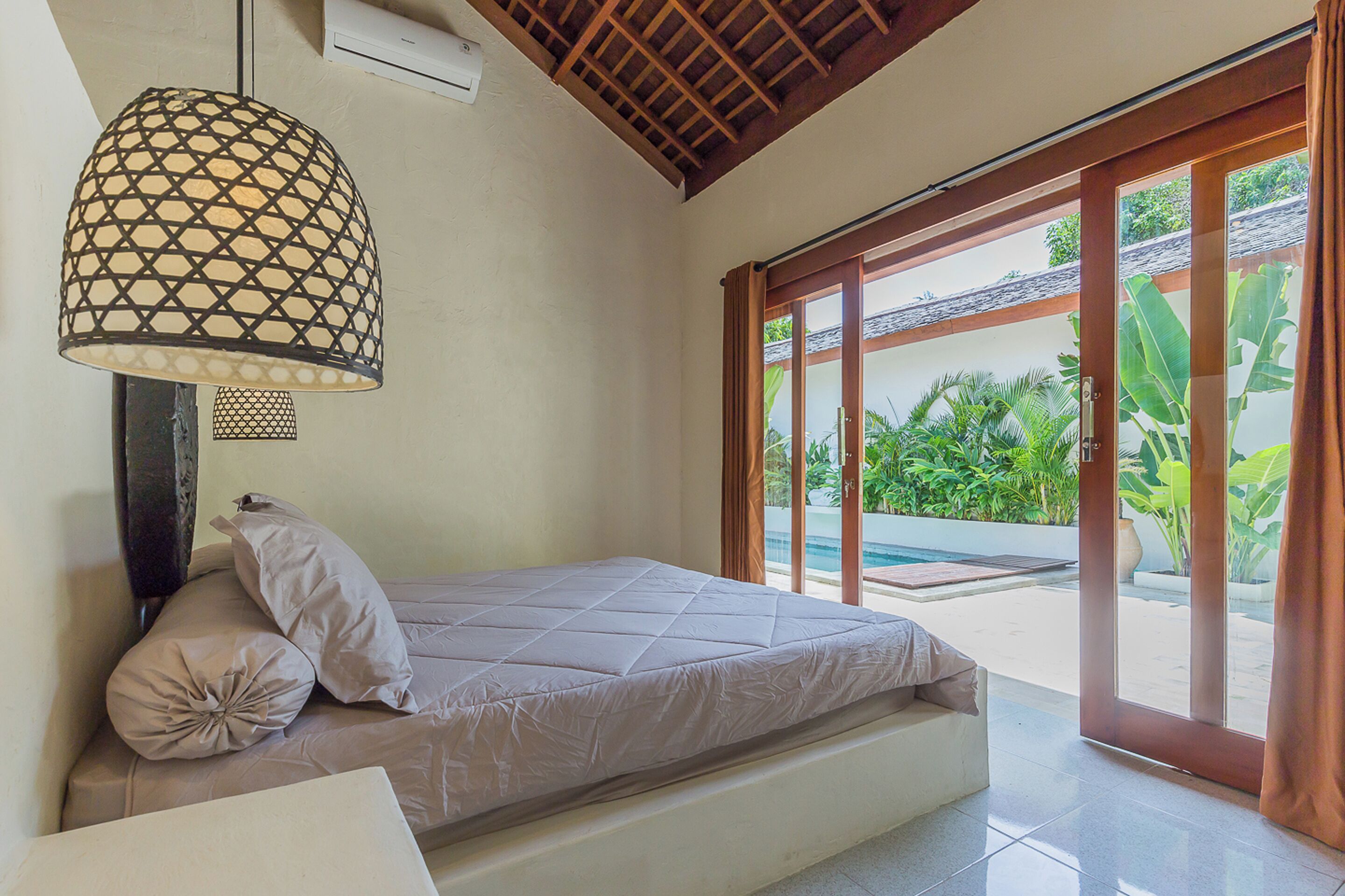 Une chambre avec un lit, une lampe suspendue et une porte-fenêtre coulissante donnant sur une zone avec piscine.