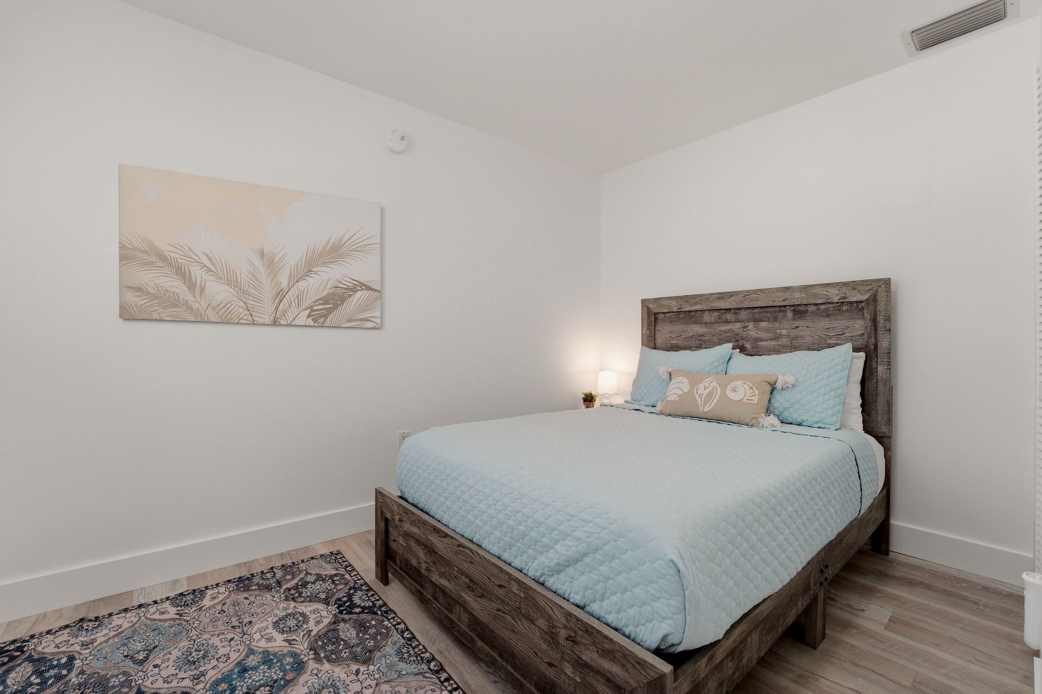Un dormitorio con estructura de cama de madera, alfombra estampada, lámpara de pared y una obra de arte enmarcada.