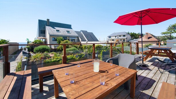 Una terraza de madera con mesas y sillas, un paraguas rojo y vistas a edificios al fondo.