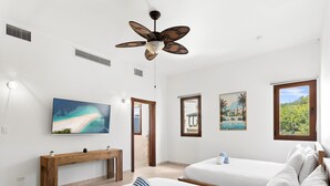 A hotel room with two beds, a ceiling fan, a painting of a beach, a small table, and a window with a view of greenery. - Stunning 2 bedroom unit Porto Cupecoy (Cupecoy)