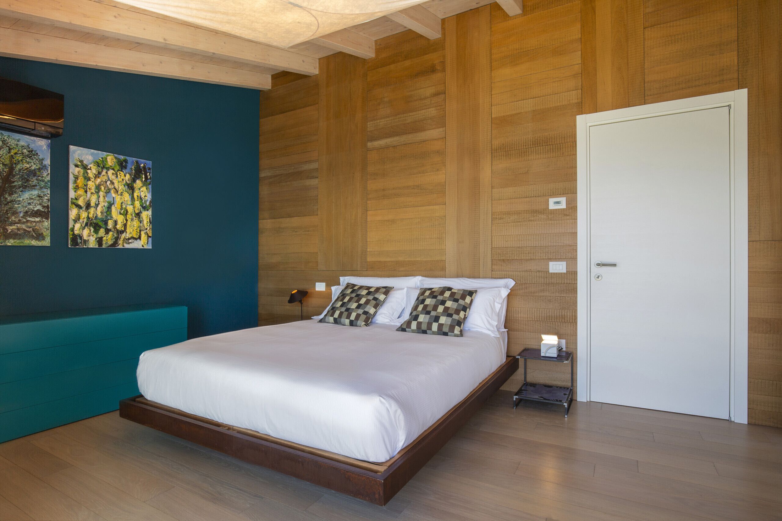 Una camera da letto con una testiera in legno, una panca color verde acqua, un letto bianco e due quadri appesi al muro.