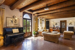 Une piĂšce avec un piano, des meubles en osier, une cheminĂ©e et des Ćuvres dâart encadrĂ©es accrochĂ©es aux murs.