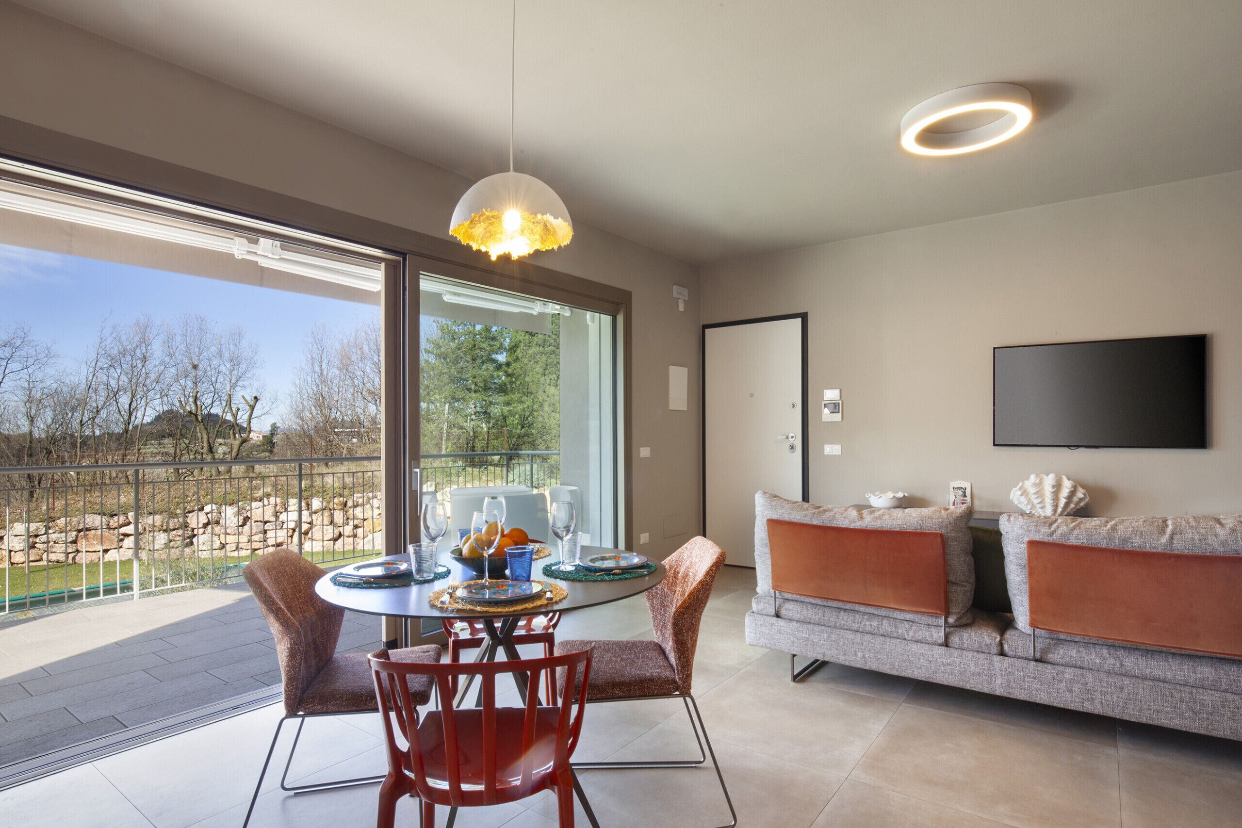 A modern living room with a dining area, a sofa, a TV mounted on the wall, and a large window offering a view of the outdoors.
