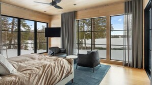 Un dormitorio moderno con una cama grande, un televisor de pantalla plana, un ventilador de techo y ventanas amplias con vistas a árboles.