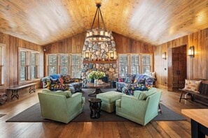 Ein geräumiger Raum mit Holzwänden und -decke, einem Steinkamin und zwei grünen Sofas mit bunten Kissen.