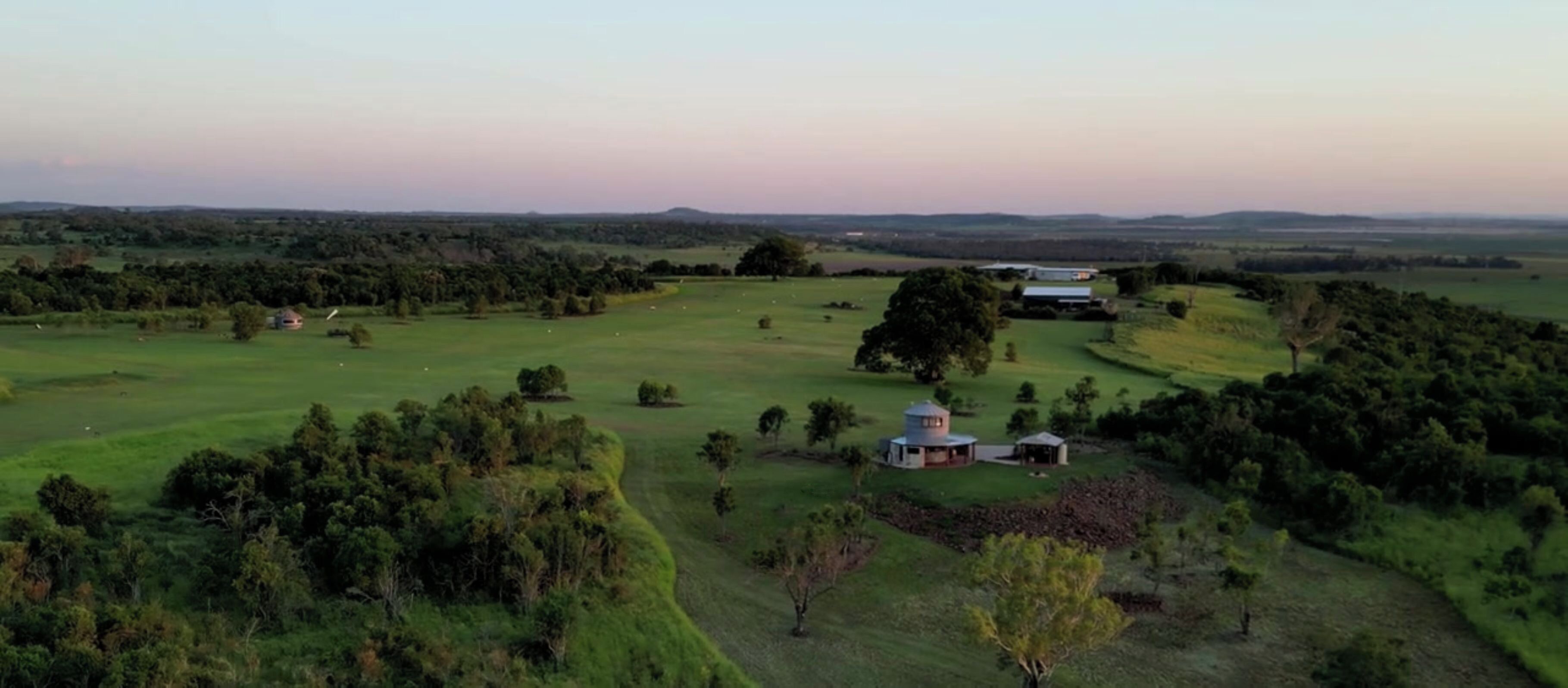 An aerial view of a rural landscape with a large open field, scattered trees, and a small building with a circular structure.