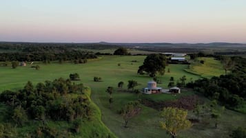 An aerial view of a rural landscape with a large open field, scattered trees, and a small building with a circular structure.