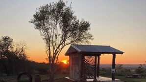 Ein Pavillon mit Metalldach, umgeben von Kies und Pflanzen, mit einem Sonnenuntergang im Hintergrund.