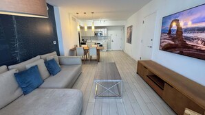 A modern living room with a sofa, a coffee table, and a TV. There is a kitchen in the background with a dining area. - 2BR High-Floor Suite · Waterway Views (Hallandale Beach)