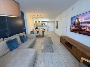 Ein modernes Wohnzimmer mit Sofa, Couchtisch und Fernseher. Im Hintergrund befindet sich eine Küche mit Essbereich.