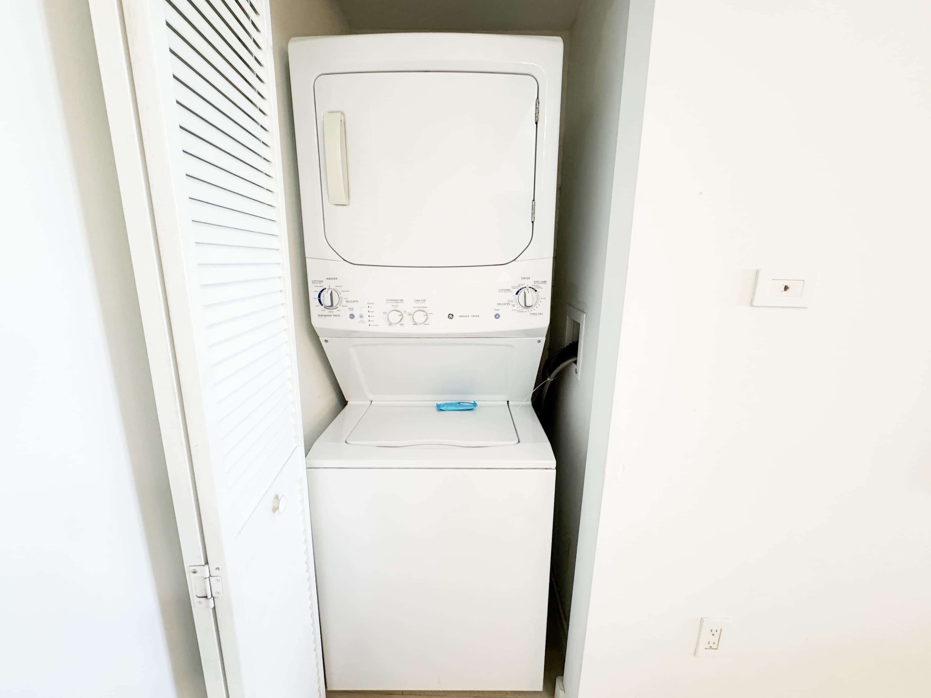 A white washing machine in a small, enclosed laundry area with a door and a wall-mounted light switch.