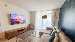Sala de estar con televisor de pantalla plana, sofá, sillón, mesa de centro y un amplio ventanal con cortinas.