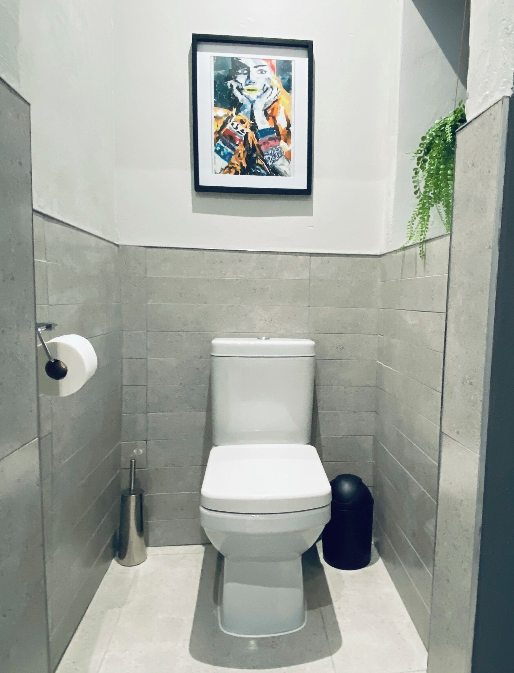 A bathroom with a toilet, a toilet paper holder, a trash bin, and a framed artwork on the wall.