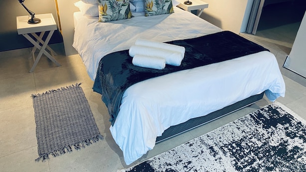 A neatly made bed with a black and white throw blanket, white pillows, and a tropical leaf-patterned pillow. There are two bedside lamps on white nightstands, a woven rug, and a black and white patterned rug on the floor. - 46 on Allcock Guest Apartment & Suite (Pretoria)