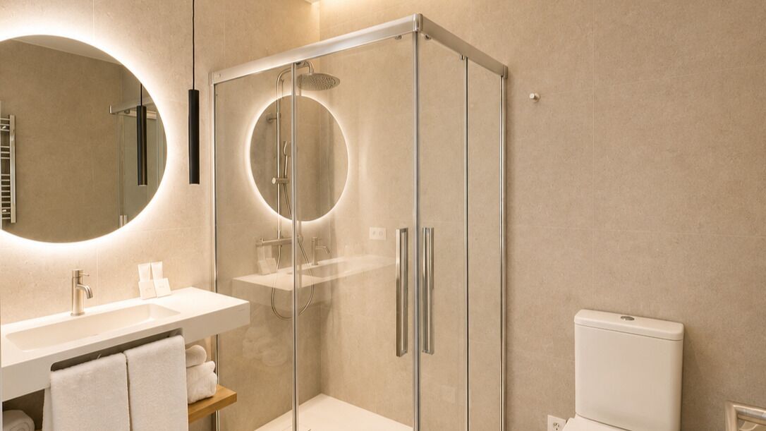 Ein modernes Badezimmer mit einer gläsernen Duschkabine, einem runden Spiegel mit Beleuchtung, einem weißen Waschbecken und einer Toilette.