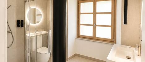 A bathroom with a glass-enclosed shower, a round mirror, a toilet, a sink, and a window with frosted glass panes.
