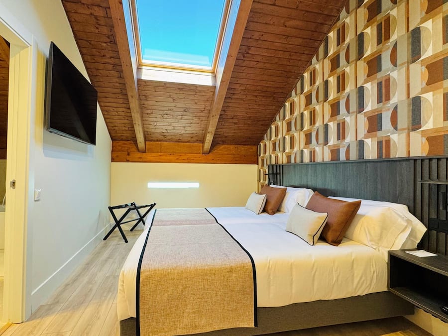 A bedroom with a sloped ceiling, a bed with pillows, a TV mounted on the wall, and a patterned wall design.