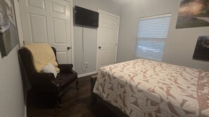 Une chambre à coucher comprenant un lit, une chaise avec une couverture, une armoire, un téléviseur et une fenêtre avec des stores.