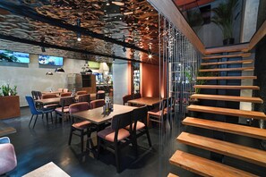 Un restaurante moderno con mesas y sillas de madera, una escalera y un área de cocina visible a través de divisiones de vidrio.
