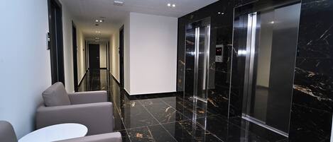 A modern hotel lobby with a marble floor, a seating area with sofas and a table, and an elevator.
