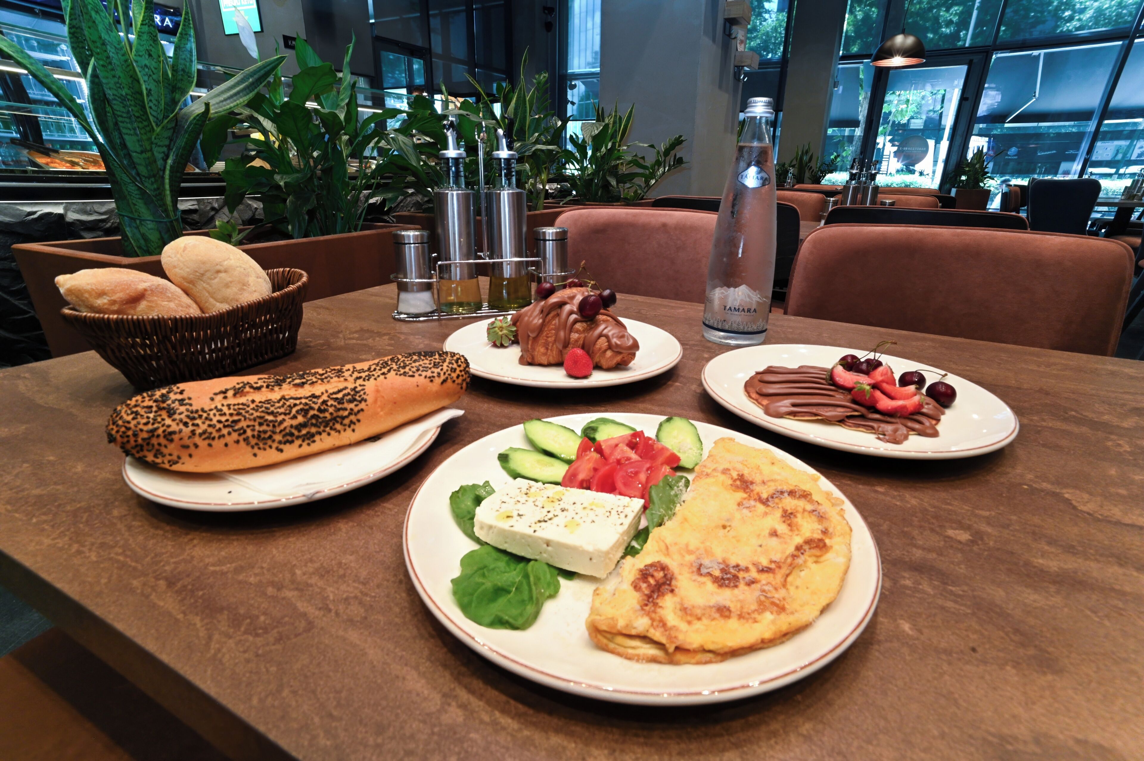 A dining table with various food items, including bread, a croissant, a plate with cheese and vegetables, and a plate with chocolate and fruit.