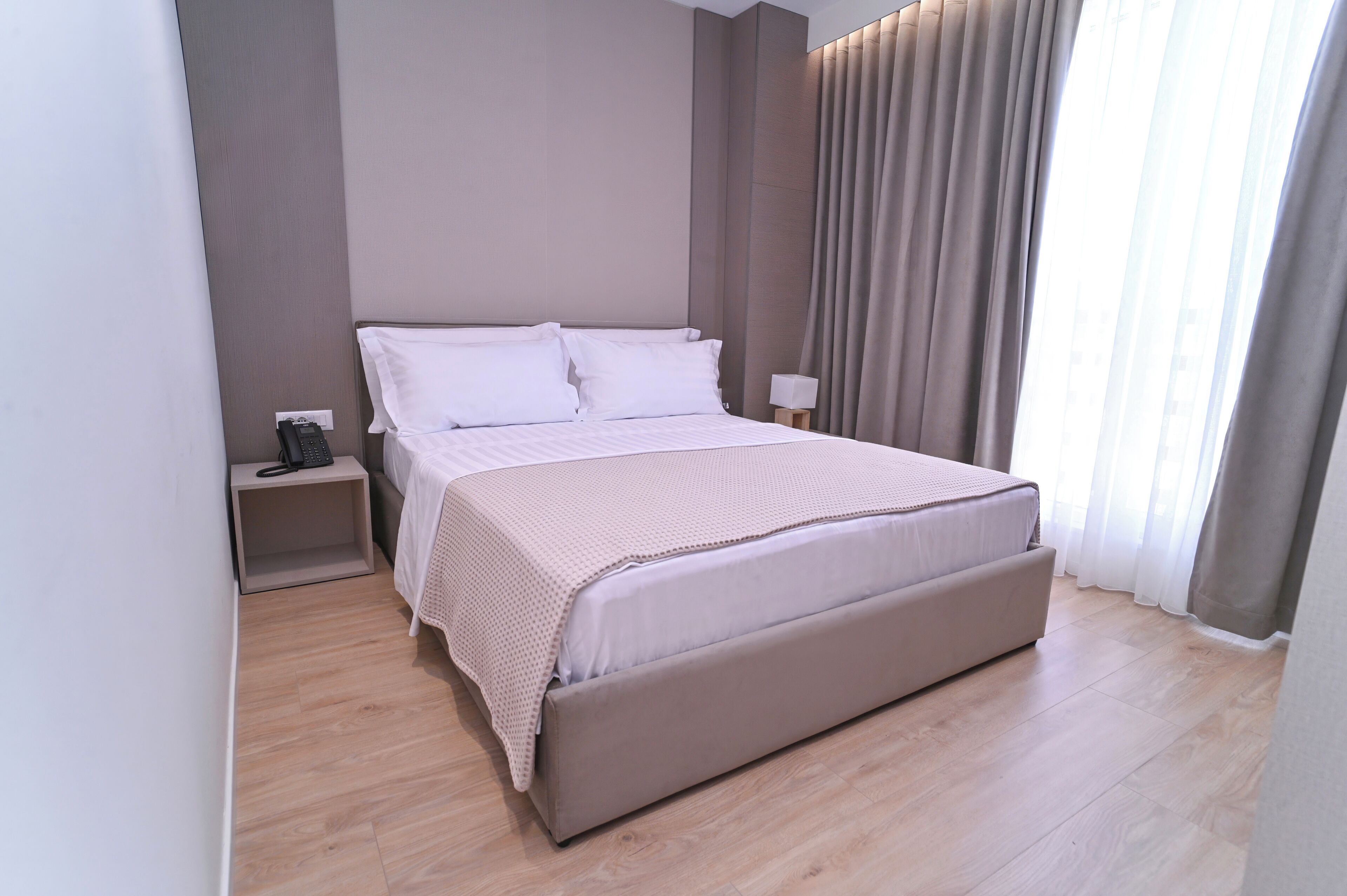 A modern hotel room with a large bed, a bedside table with a lamp, and a view of the outside through sheer curtains.