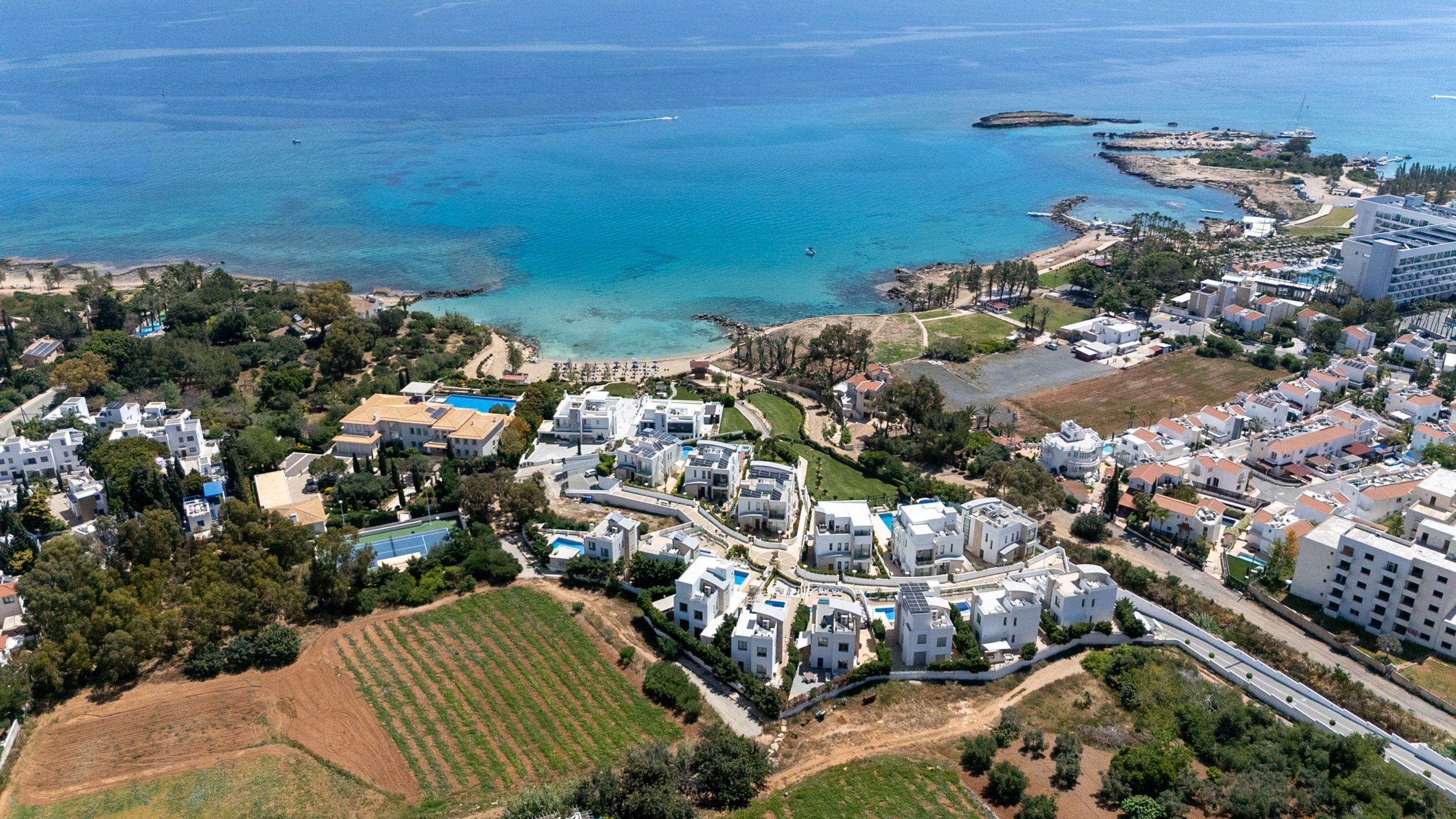 An aerial view of a coastal residential area with modern white buildings, a beach, and clear blue waters.