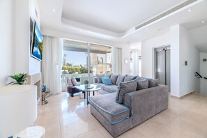 A modern living room with a grey sectional sofa, a small round table, and a large window offering a view of the outdoors. - Villa Lombardi 9 in Protaras (Paralimni)