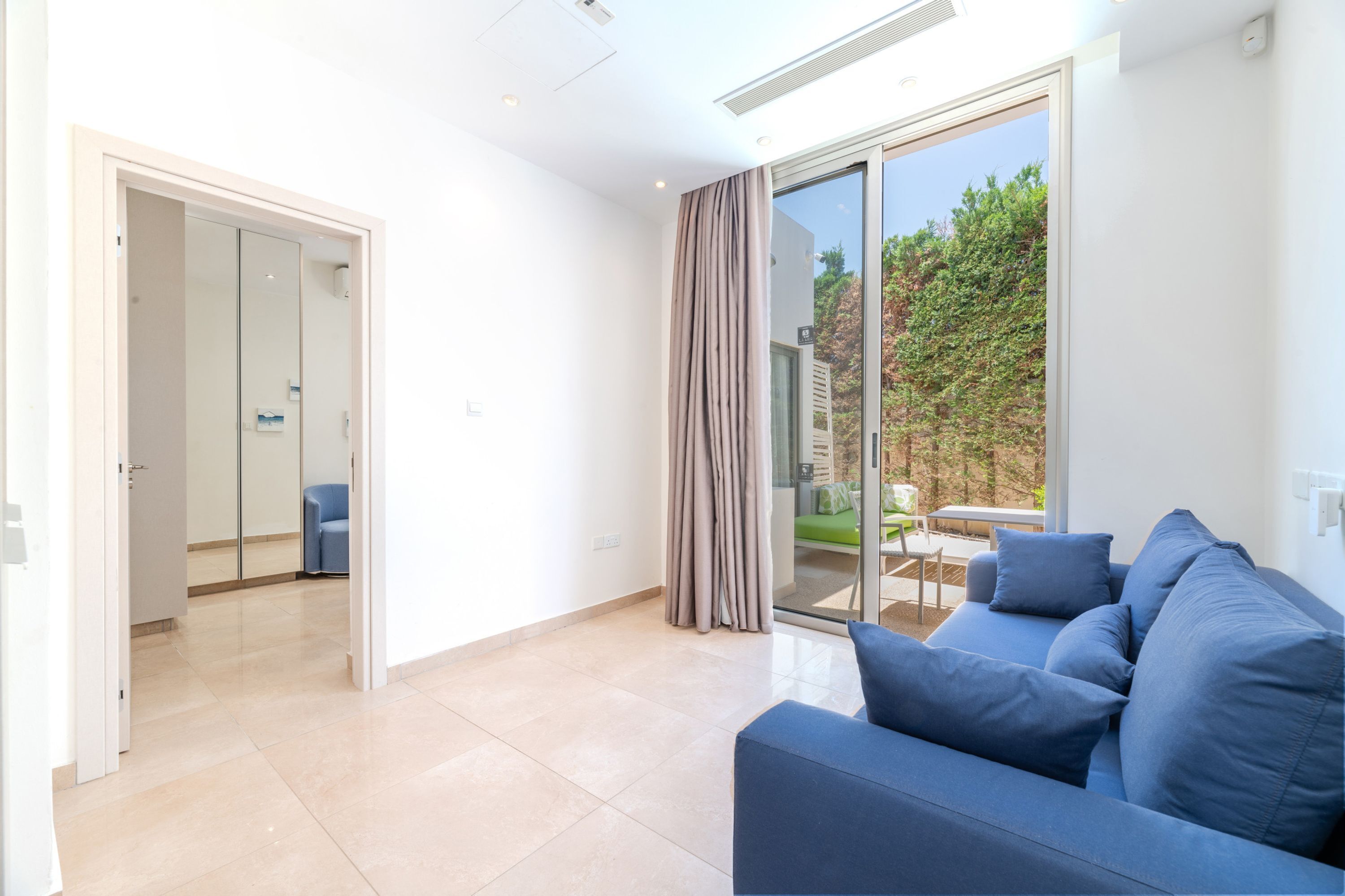 A modern living room with a blue sofa, a glass door leading to a balcony, and a view of greenery outside.