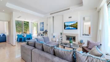 Ein modernes Wohnzimmer mit einem großen grauen Sofa, einem Essbereich mit blauen Stühlen, einem Kamin und einem großen Fenster mit Blick auf Grünflächen.