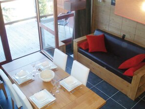 Ein Essbereich mit einem Holztisch, gedeckt für vier Personen, einer Couch mit roten Kissen und Blick durch eine Glastür auf eine Terrassenfläche.