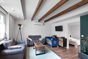 Una sala moderna con un sofá, una mesita de centro, un corralito azul, un televisor y una cama al fondo.