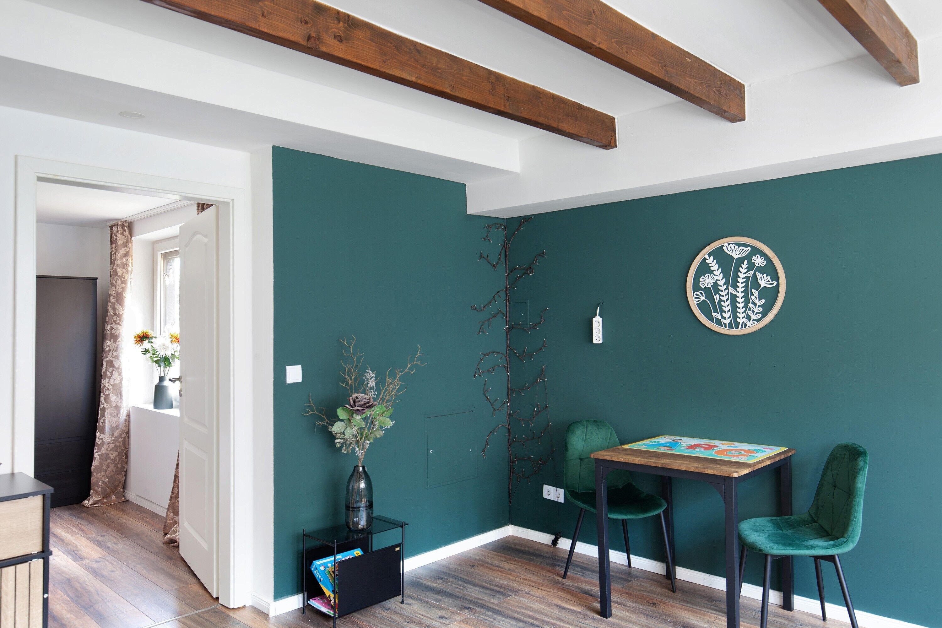 Une pièce aux murs de couleur turquoise, avec une poutre en bois au plafond, une table entourée de chaises, un vase rempli de fleurs et un élément décoratif circulaire fixé au mur.