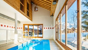 An indoor swimming pool with wooden beams, large windows, and a view of snowy houses. - Samoens Village Apartment for 4 (Samoëns)