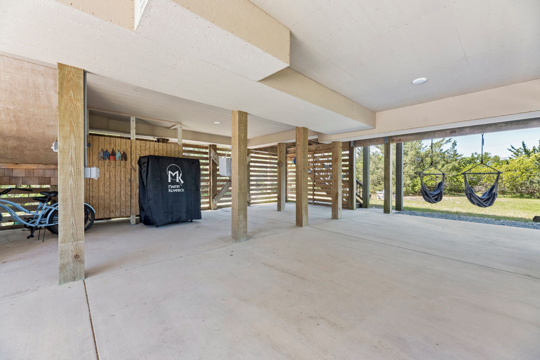 An open covered area with wooden beams, a concrete floor, and a view of greenery outside.