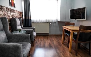 Ein Wohnzimmer mit einer Couch, einem kleinen runden Tisch, einem Holzessttisch mit Stühlen, einem an der Wand montierten Fernseher und einer Heizung.