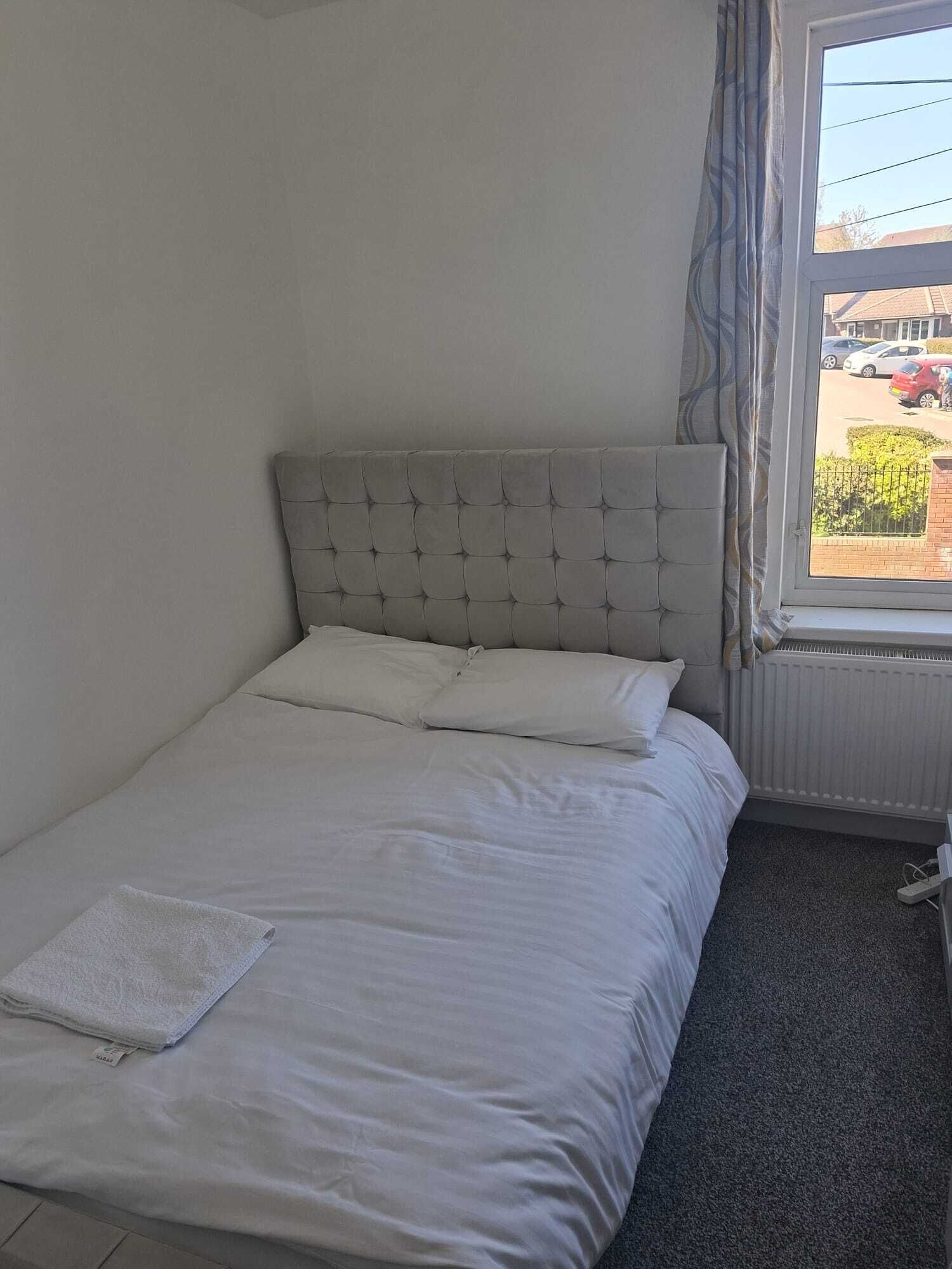 A single bed with a tufted headboard, white bedding, and a window with a view of parked cars and a building.