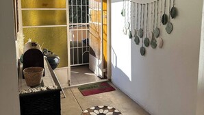 Un pasillo con suelo de baldosas, un felpudo, una cesta, un radiador y una puerta que da a una habitación con ventana.