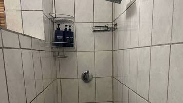 Une cabine de douche carrelée, dotée d’un pommeau de douche à main, d’une étagère fixée au mur pour les articles de toilette et d’un drain dans le sol.