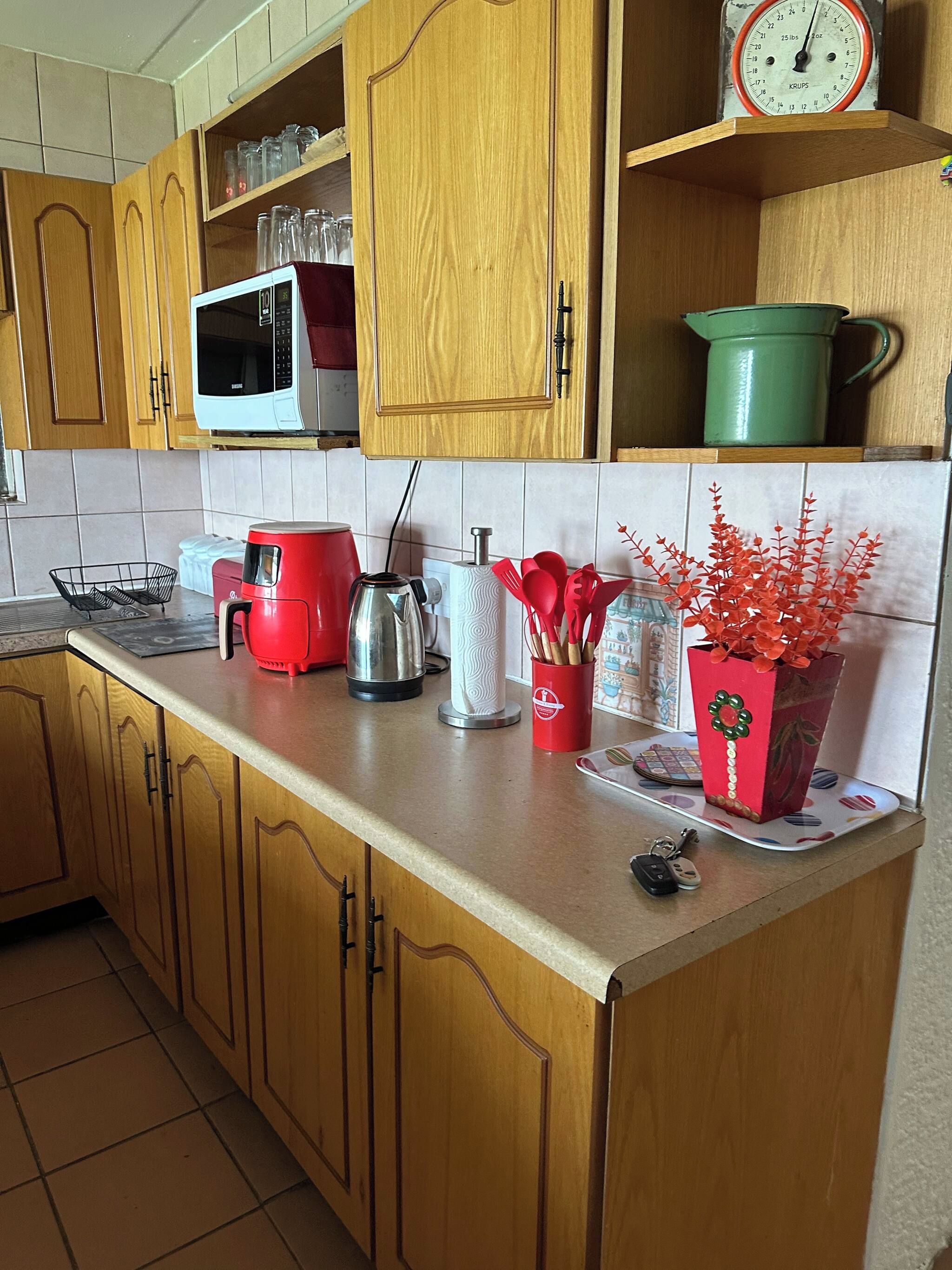 A kitchen with wooden cabinets, a microwave, a kettle, a red coffee maker, a green pot, and a decorative red vase with red flowers.